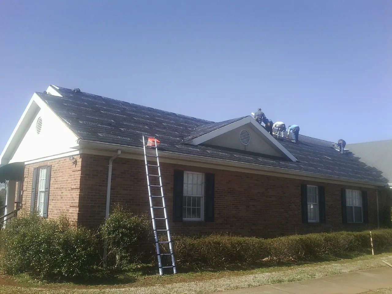 Crew tearing shingles off a brick ranch roof with a ladder set — Metal Roof Repair work by our crew in Stevens