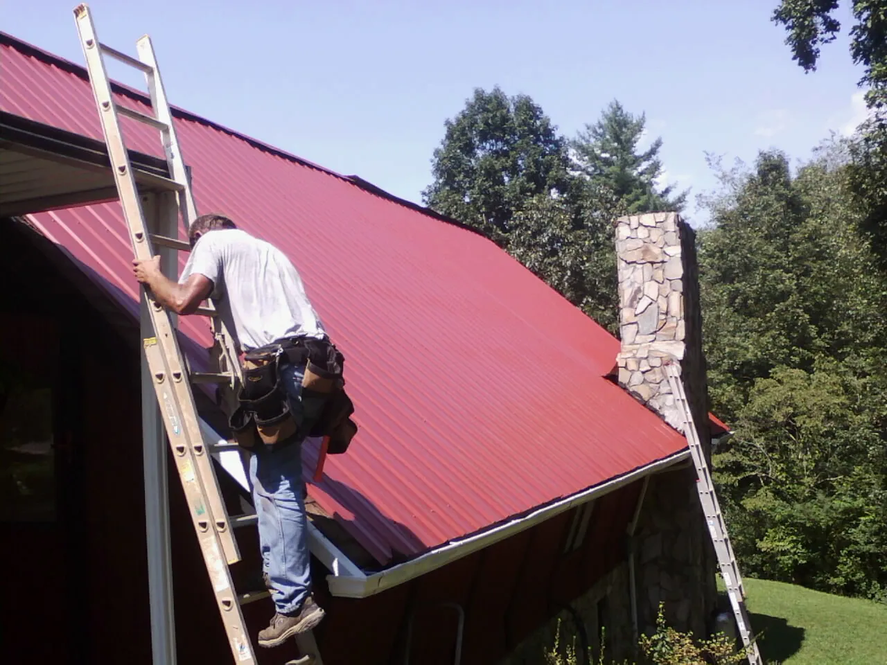 Roofer on a ladder beside a red metal roof and stone chimney — Soffit & Fascia Repair by Roofing Brothers in Stevens